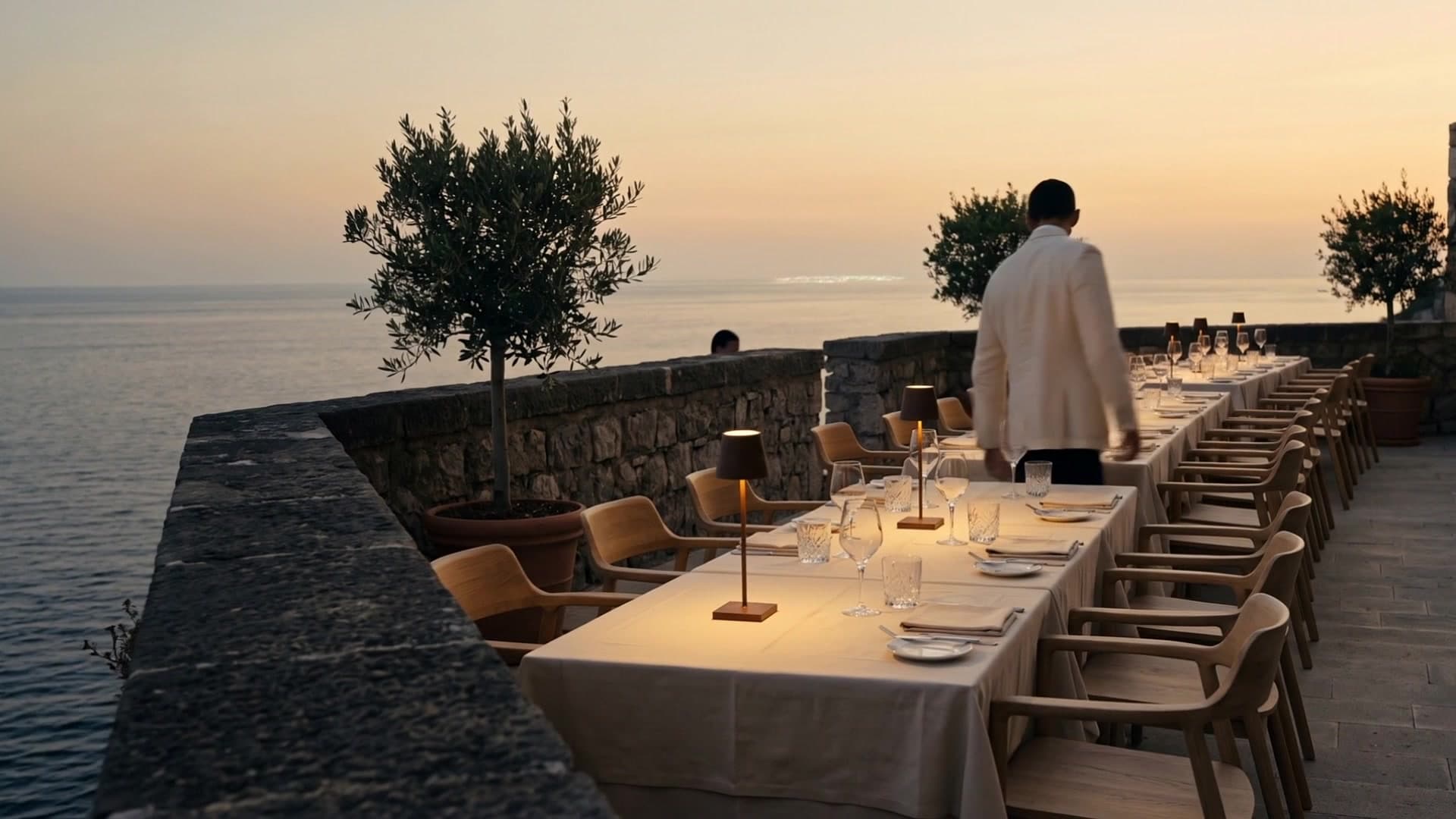 At dusk, a maître d’hôtel checks a long set table on a terrace above the sea